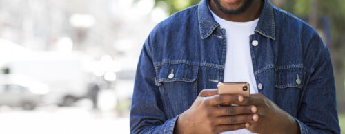 Homem negro, de camiseta branca e jaqueta jeans, manuseando telefone celular ao ar livre