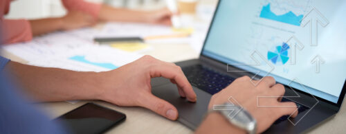 Mãos de homem digitando em notebook em ambiente de trabalho, com pessoa trabalhando ao fundo