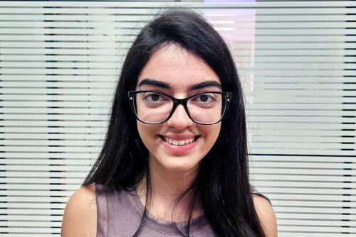 Mulher de 19 anos, sorrindo. Cabelo preto, óculos de grau e blusa estilo regata.