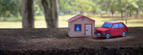 Carro e casa de papel, um do lado do outro, no chão e com árvores no fundo.
