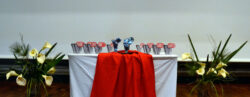 Foto de uma mesa com um tecido branco e um vermelho estendidos. Sobre a mesa estão os troféus. Aos dois lados da mesa, estão dois vasos de flores