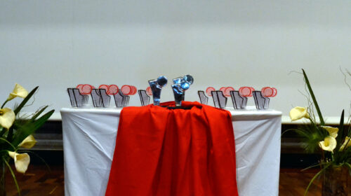 Foto de uma mesa com um tecido branco e um vermelho estendidos. Sobre a mesa estão os troféus. Aos dois lados da mesa, estão dois vasos de flores